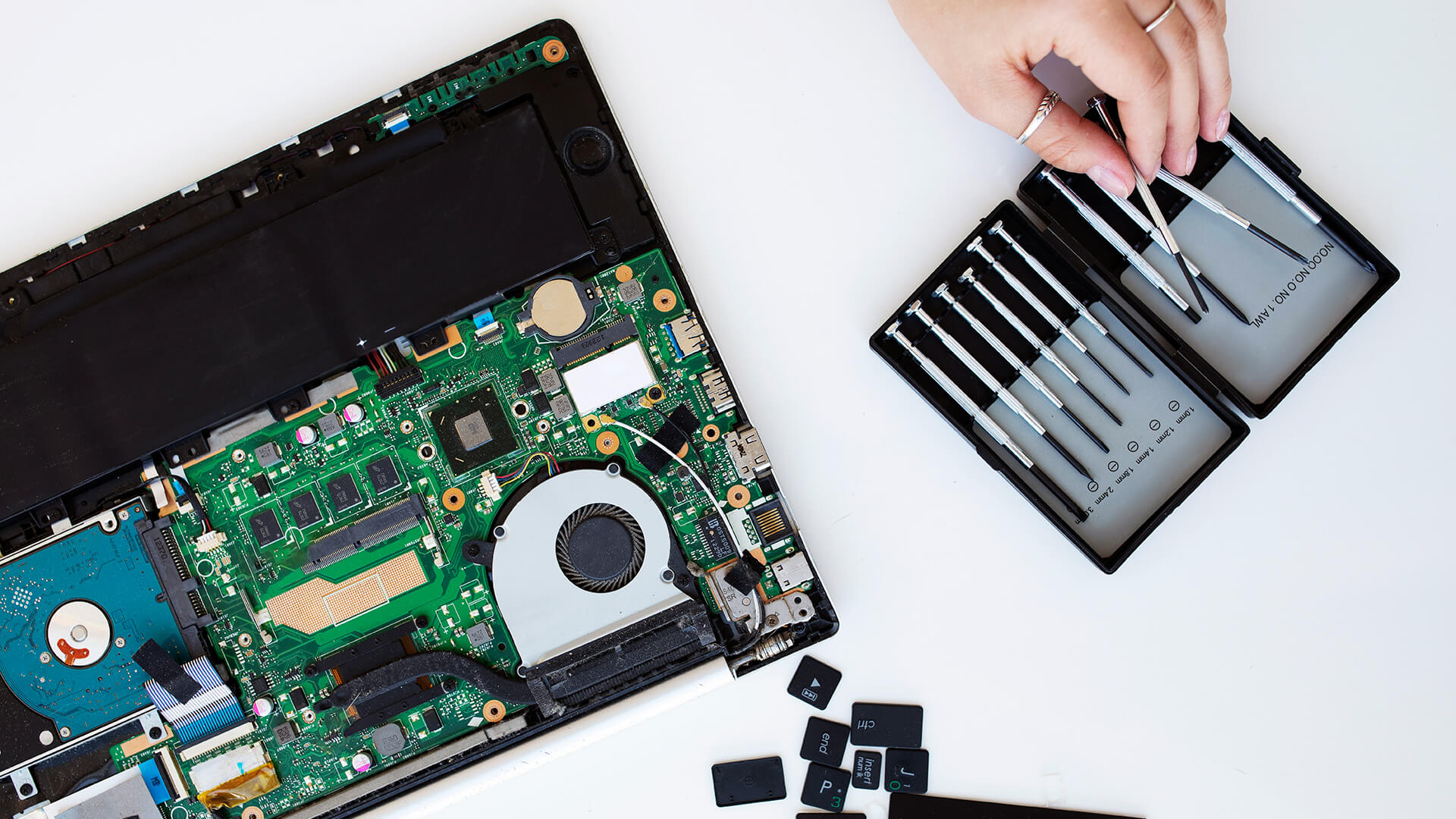 Pessoa realizando manutenção em um laptop com ferramentas específicas, com a tampa do dispositivo aberta revelando a placa-mãe, na frente de uma caixa de utensílios para reparo de eletrônicos.