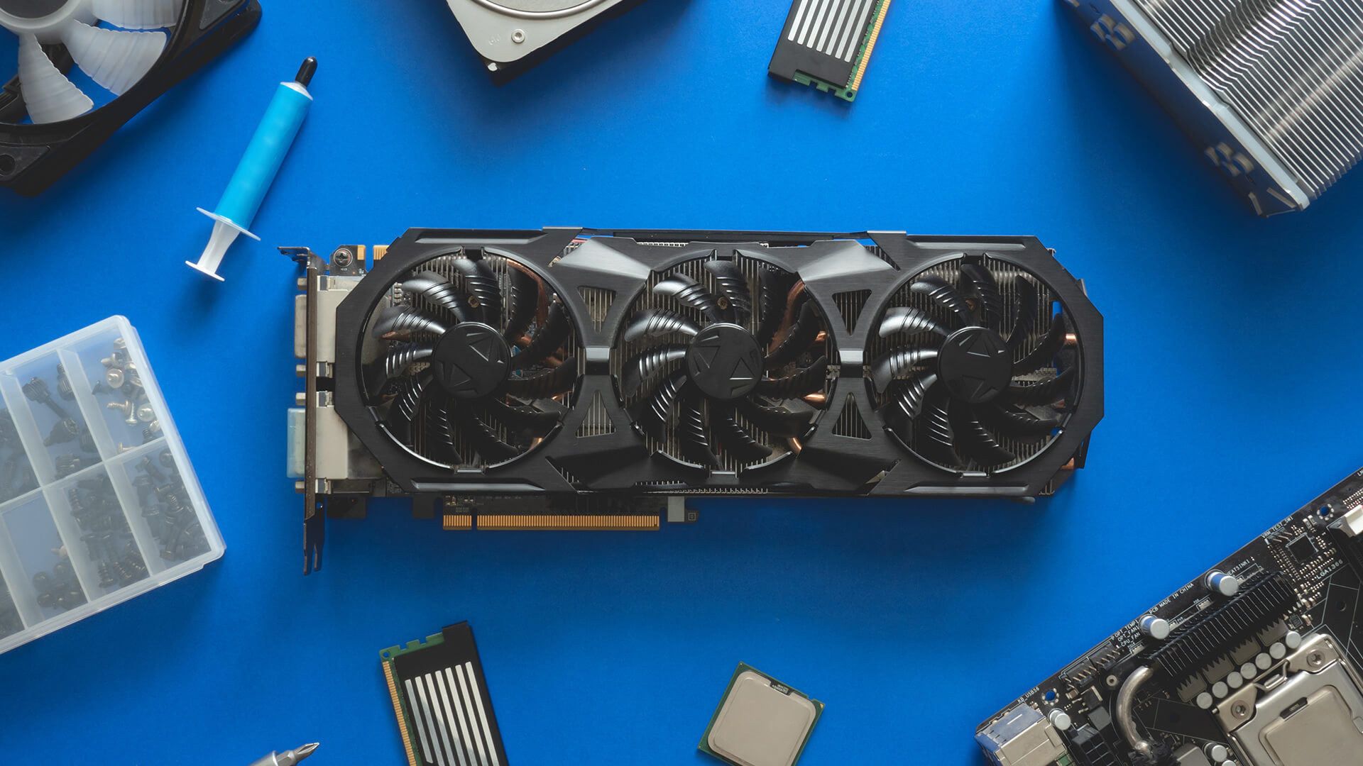Placa de v&iacute;deo GTX com tr&ecirc;s ventoinhas em bancada de montagem de computadores, acompanhada de componentes de hardware e ferramentas.